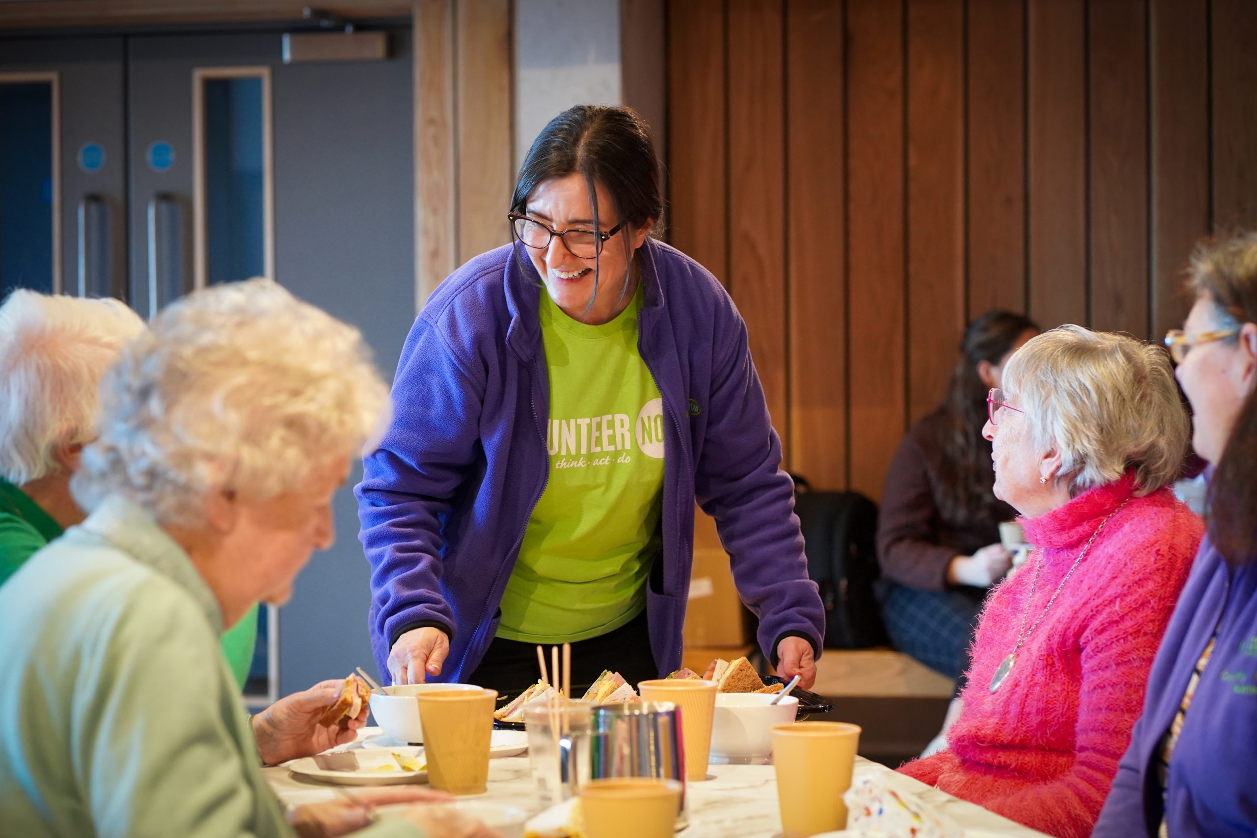 Smiling woman in a lime green Volunteer Now tshirt and purple fleece serving a table of women at an older persons group get together