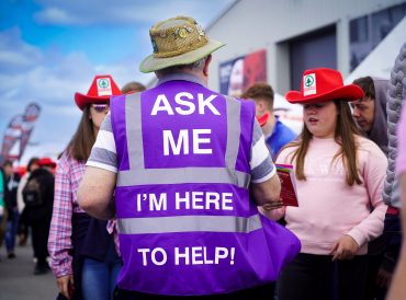Volunteer with his back to the camera in a crowd, wearing a purple high viz vest with Ask me I'm here to help on the back.