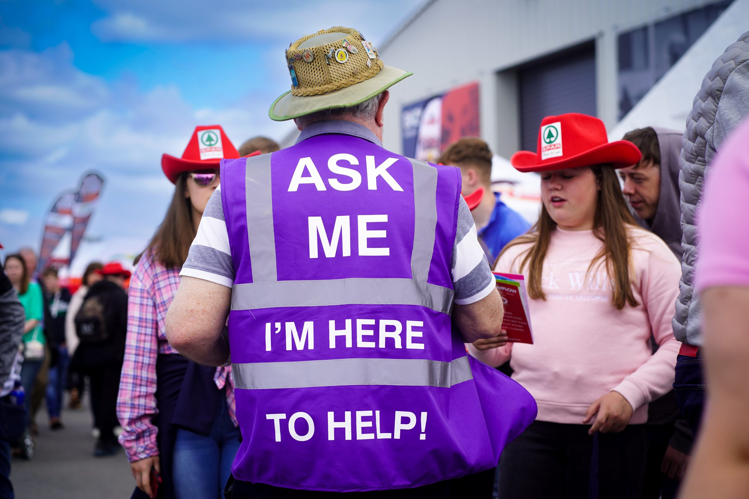 Volunteer with his back to the camera in a crowd, wearing a purple high viz vest with Ask me I'm here to help on the back.