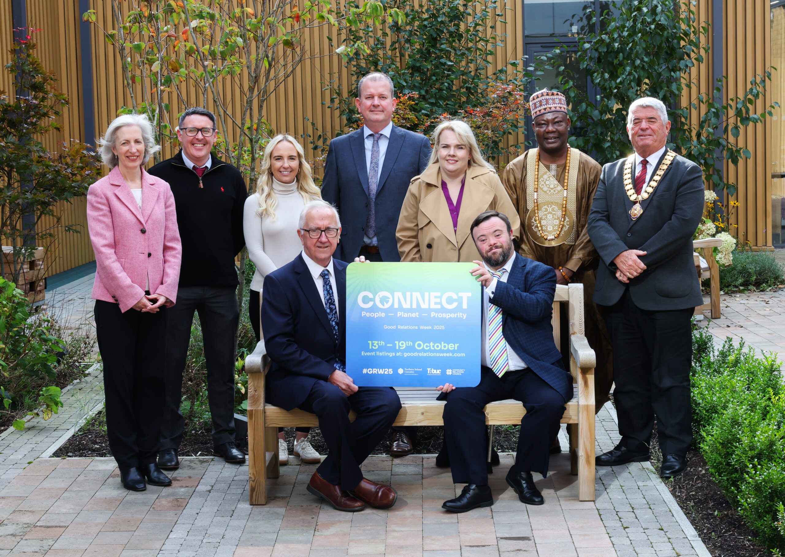 Photo of a group of people smiling - 7 standing behind 2 people sitting holding a sign that says CONNECT people - planet - prosperity for Good Relations Week 2025