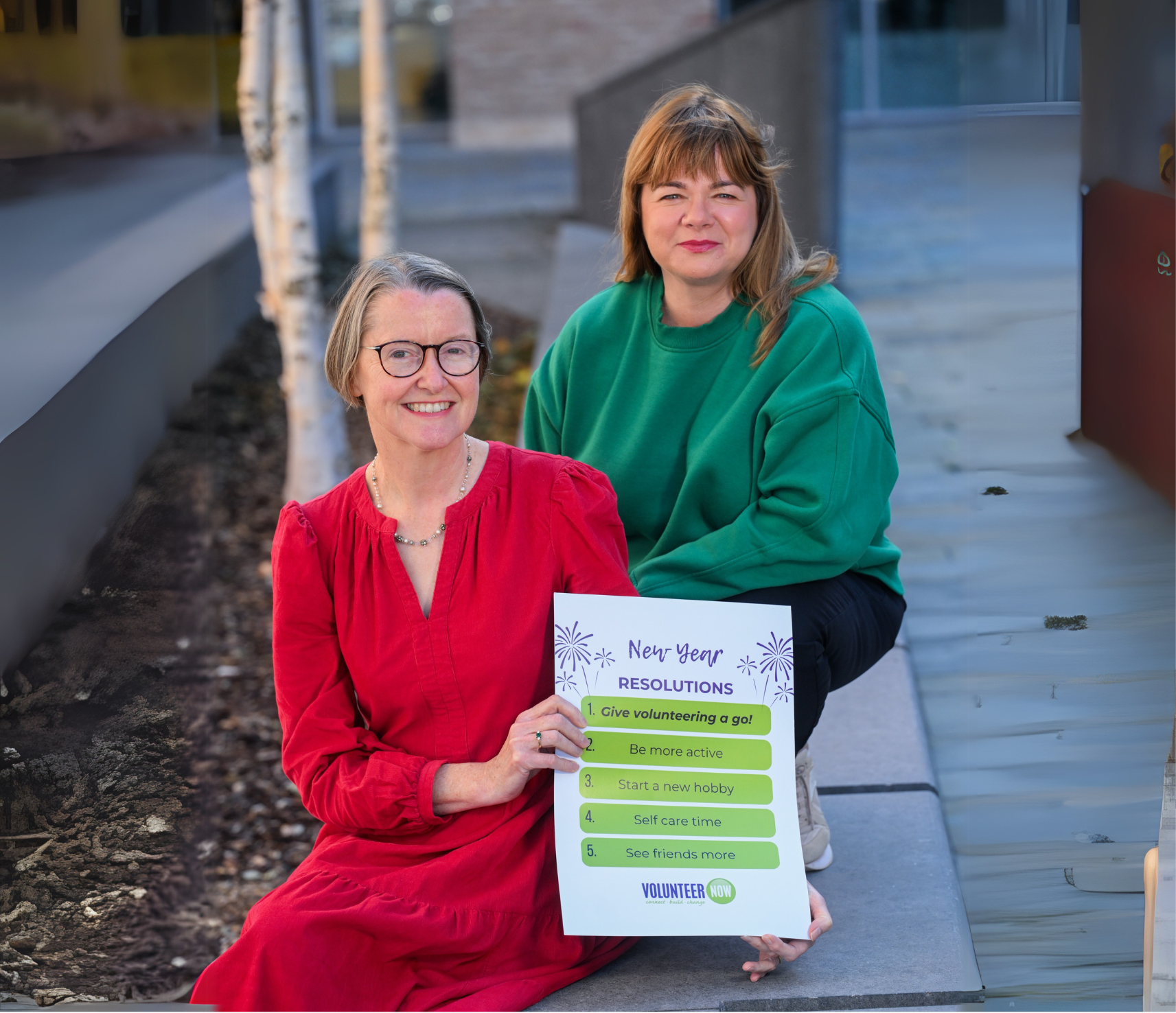 Two smiling women, one in a red dress, the other wiearing a green jumper holding a sign for New Years resolutions