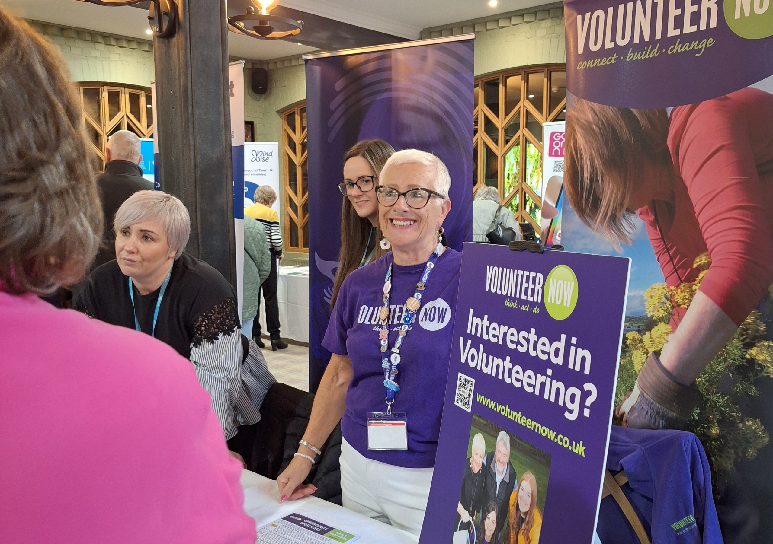 Wenda, our Volunteering Support Officer, at an information stand beside a poster that says Interested in Volunteering?