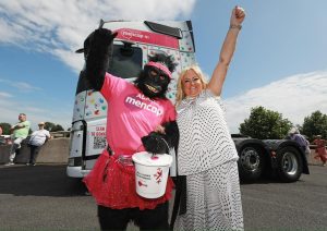 A woman stands beside someone dressed as a gorilla with a Mencap pink t-shirt and red tutu, and holding a collection bucket. Both have an arm in the air and in the background is a big lorry cab with Mencap branding.