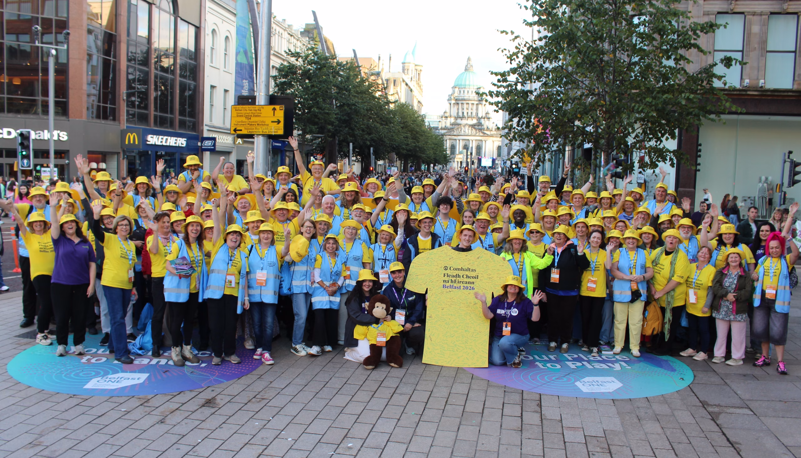 A group of 100+ Fleadh 2026 Event Volunteers, all wearing their bright yellow t-shirts and smiling and waving at the camera